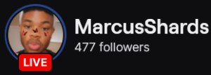 MarcusShards' Twitch logo and follower count (477). Logo is a picture of a black man with a lowcut afro and red/black butterfly stickers on his face. Image links to MarcusShards' Twitch page.